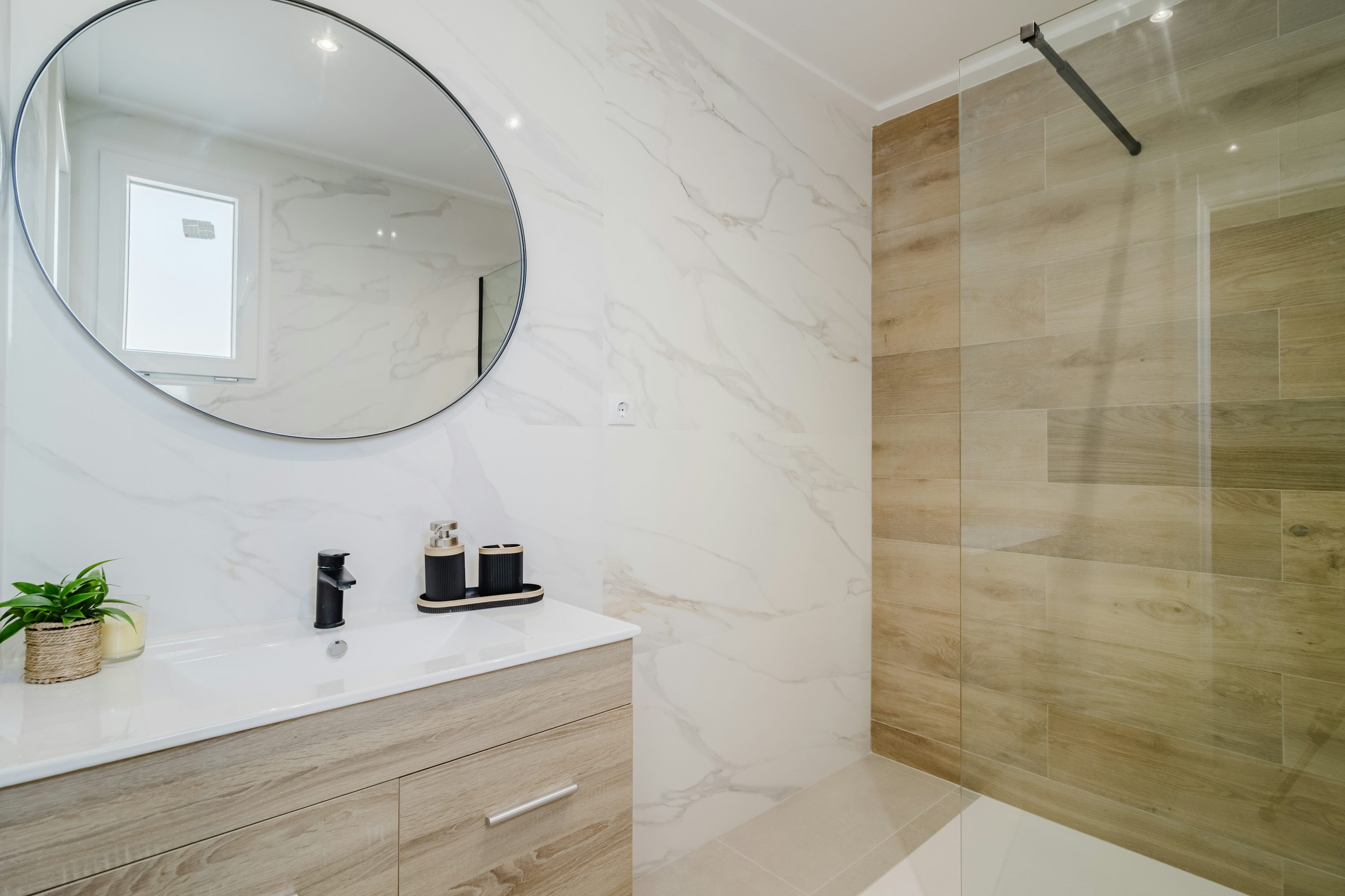 Modern bathroom with a wooden and marble design featuring a round mirror and glass shower.