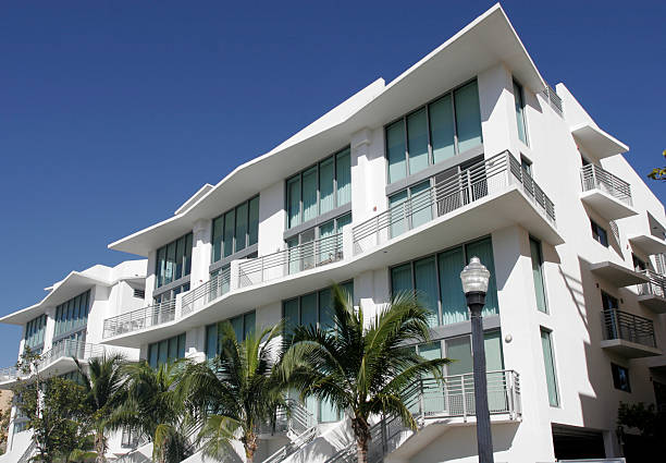 Modern white condominium building with large windows and palm trees under a blue sky.